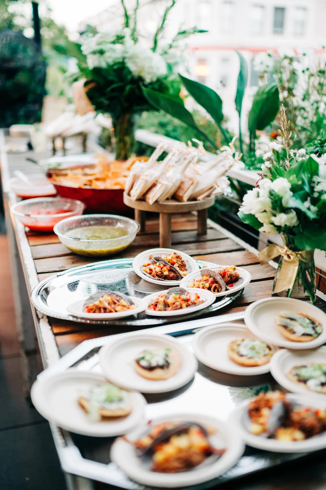Private event catering table at Darling Aviary Sacramento with mini pulled-pork tacos on blue corn tortillas, tostadas, tortilla chips, red salsa, salsa verde, and lush white floral arrangements on a K Street rooftop