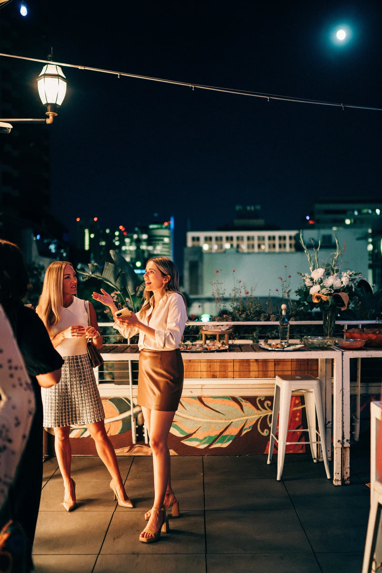 Two women in evening attire chatting on the Darling Aviary rooftop at night with the Downtown Sacramento skyline, full moon, string lights, vintage lantern, and a styled bar with floral arrangements — K Street rooftop event