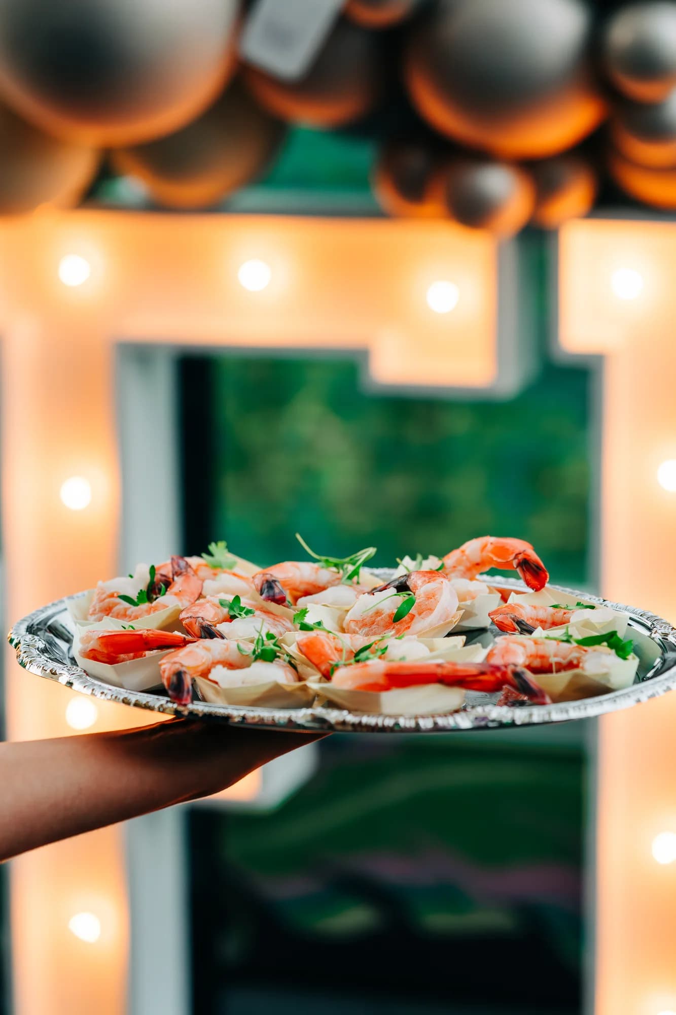 Silver platter of chilled jumbo shrimp cocktail in individual paper cups with fresh microgreens, served at a Darling Aviary private event on the Sacramento rooftop with a lit marquee letter and balloon backdrop