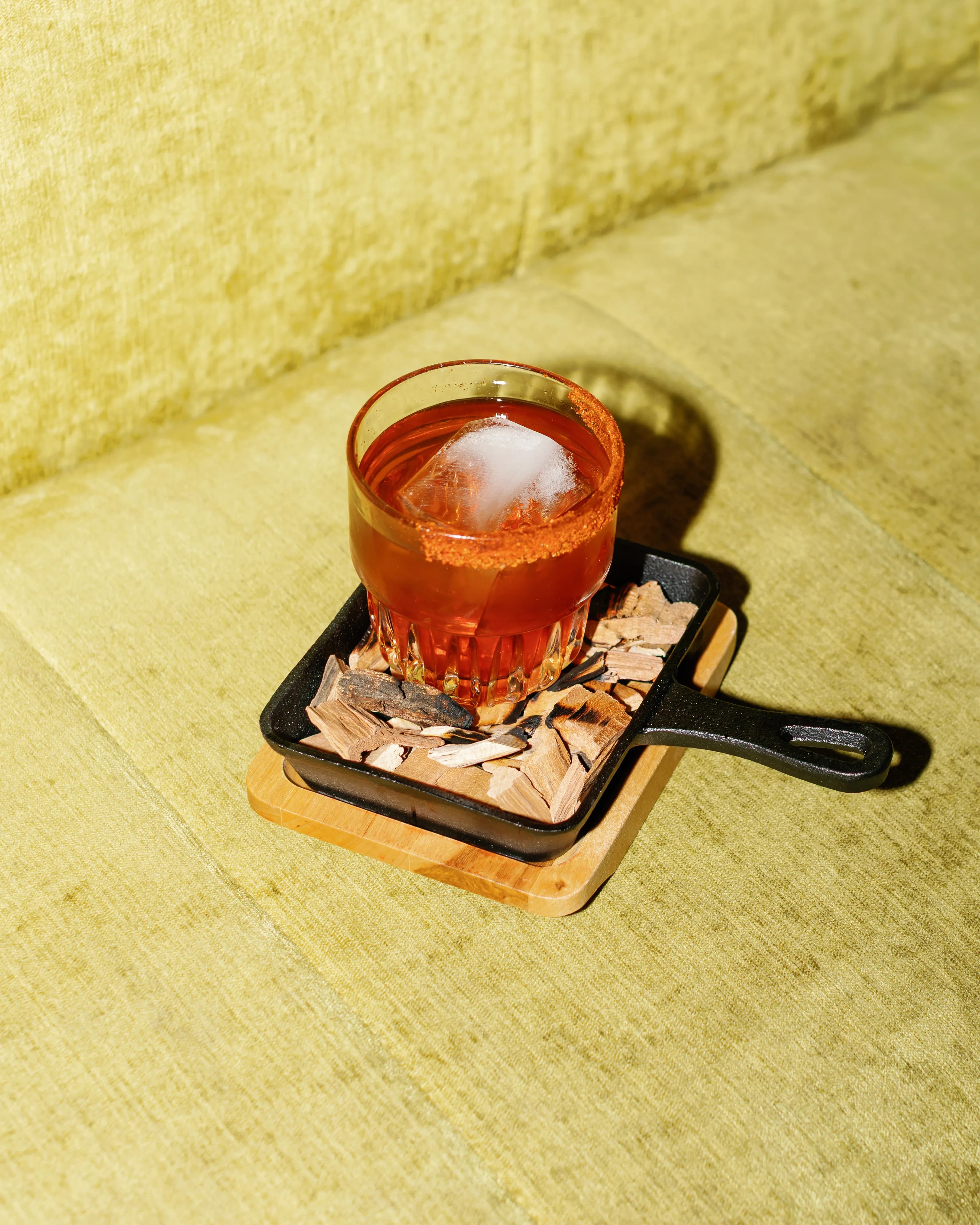 Darling Aviary Smoked Old Fashioned in a rocks glass on a cast iron skillet with wood chips, served on a chartreuse velvet sofa at rooftop cocktail bar in DoCo, Downtown Sacramento
